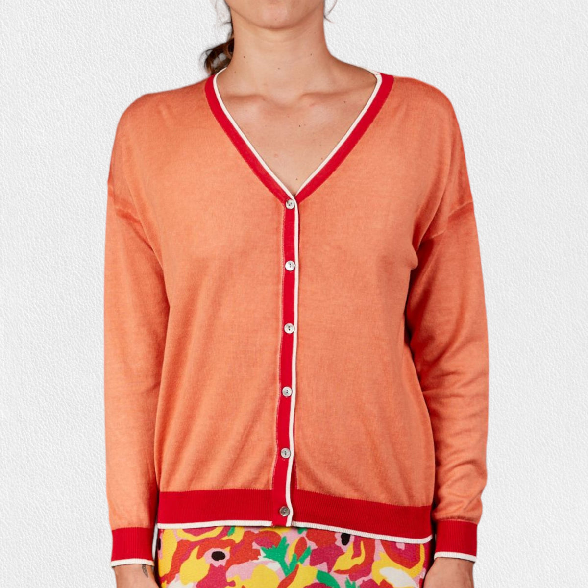 A person is wearing a vibrant coral-colored IN BED WITH YOU - BATWING SLEEVE CARDIGAN by IN BED WITH YOU/CLAUDIOLAI SRL, featuring red and white trim and buttoned down the front. The bottom hem and cuffs are solid red. They also have on bright, floral-patterned shorts. The person&#39;s face is not visible. This ensemble epitomizes the signature look of a distinguished Italian brand.