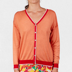 A woman wears the IN BED WITH YOU - BATWING SLEEVE CARDIGAN by IN BED WITH YOU/CLAUDIOLAI SRL, an orange V-neck with red and white trim, buttoned down the front, styled with multicolored patterned pants against a plain white background.