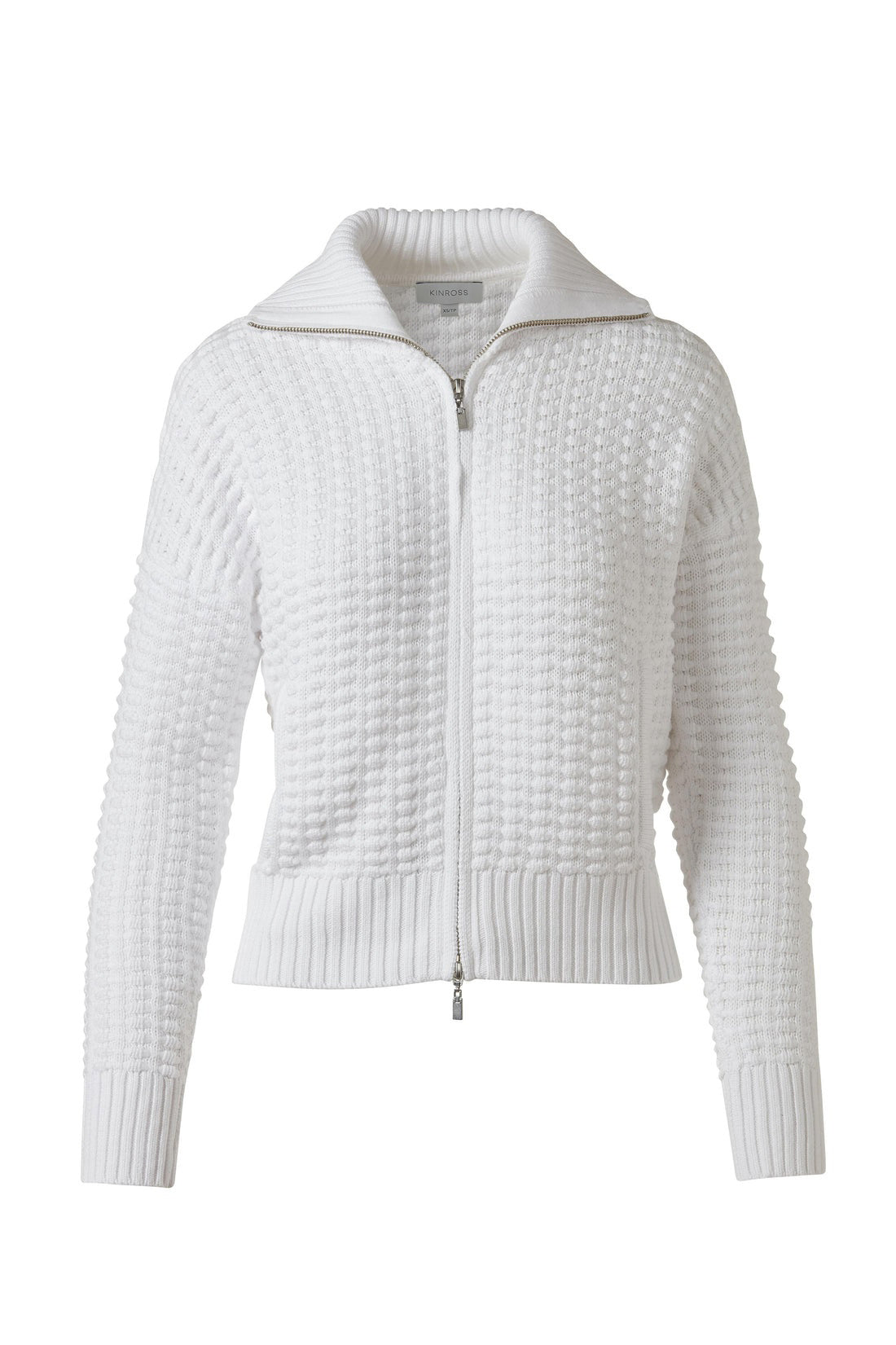 A woman wears the KINROSS Bobble Zip Cotton Cardigan in white over a black and beige striped button-up shirt with white pants, standing facing forward with one hand in her pocket against a plain white background.