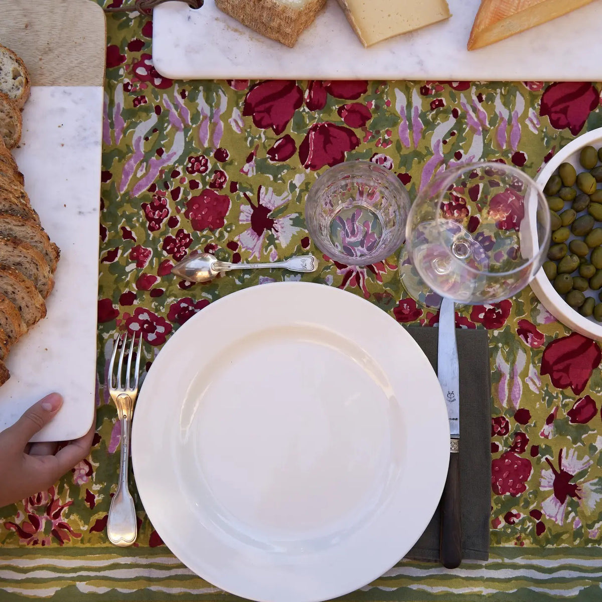 On the terrace, a blue chair holds a straw hat next to a table adorned with COULEUR NATURE&#39;s JARDIN RED AND GREEN TABLECLOTH 59X59. Two glasses of red wine and a wine bottle set the scene against lush trees and rolling hills, crafting an idyllic countryside backdrop.
