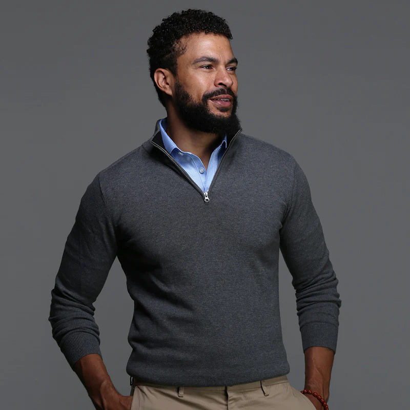 A man with short dark hair and a trimmed beard wears the COLLARS AND CO - SEMI SPREAD COLLAR POLO BLUE HOUNDSTOOTH, paired with light-colored pants. He stands indoors, leaning against a column with a neutral expression.