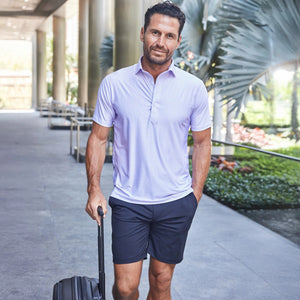A man with short dark hair and a beard, wearing the COLLARS AND CO - Featherweight Flex Polo in Vantage Lavender with a dress collar and 6-way stretch, stands outdoors before large green palm leaves, looking to the side.