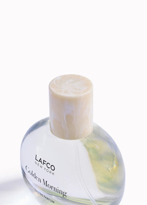 A transparent perfume bottle labeled “LAFCO - FINE FRAGRANCE, GOLDEN MORNING 50ML” by LAFCO features a round, marble-textured cream cap and is displayed against a plain white background. This floral scent is crafted as a vegan and cruelty-free fine fragrance, designed to deliver a sophisticated and conscious olfactory experience.