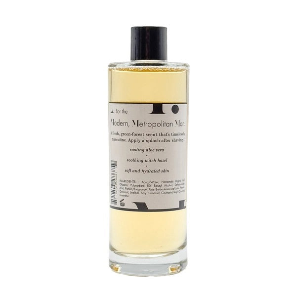 A clear bottle filled with yellow liquid, BAUDELAIRE - M.B. AFTERSHAVE by BAUDELAIRE features a classic design with a black hat graphic on the front and a black cap, offering a refreshing green-forest scent enriched with soothing witch hazel.