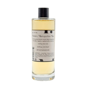 A clear bottle filled with yellow liquid, BAUDELAIRE - M.B. AFTERSHAVE by BAUDELAIRE features a classic design with a black hat graphic on the front and a black cap, offering a refreshing green-forest scent enriched with soothing witch hazel.