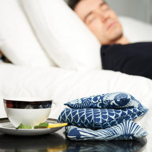 Three square pillows in blue and white patterns—a geometric wave, a radial fan motif, and circular flower-like shapes adorned with leafy details—ideally complement the use of BAUDELAIRE's Chamomile Sleep Sachets by infusing relaxation into your sleep sanctuary.