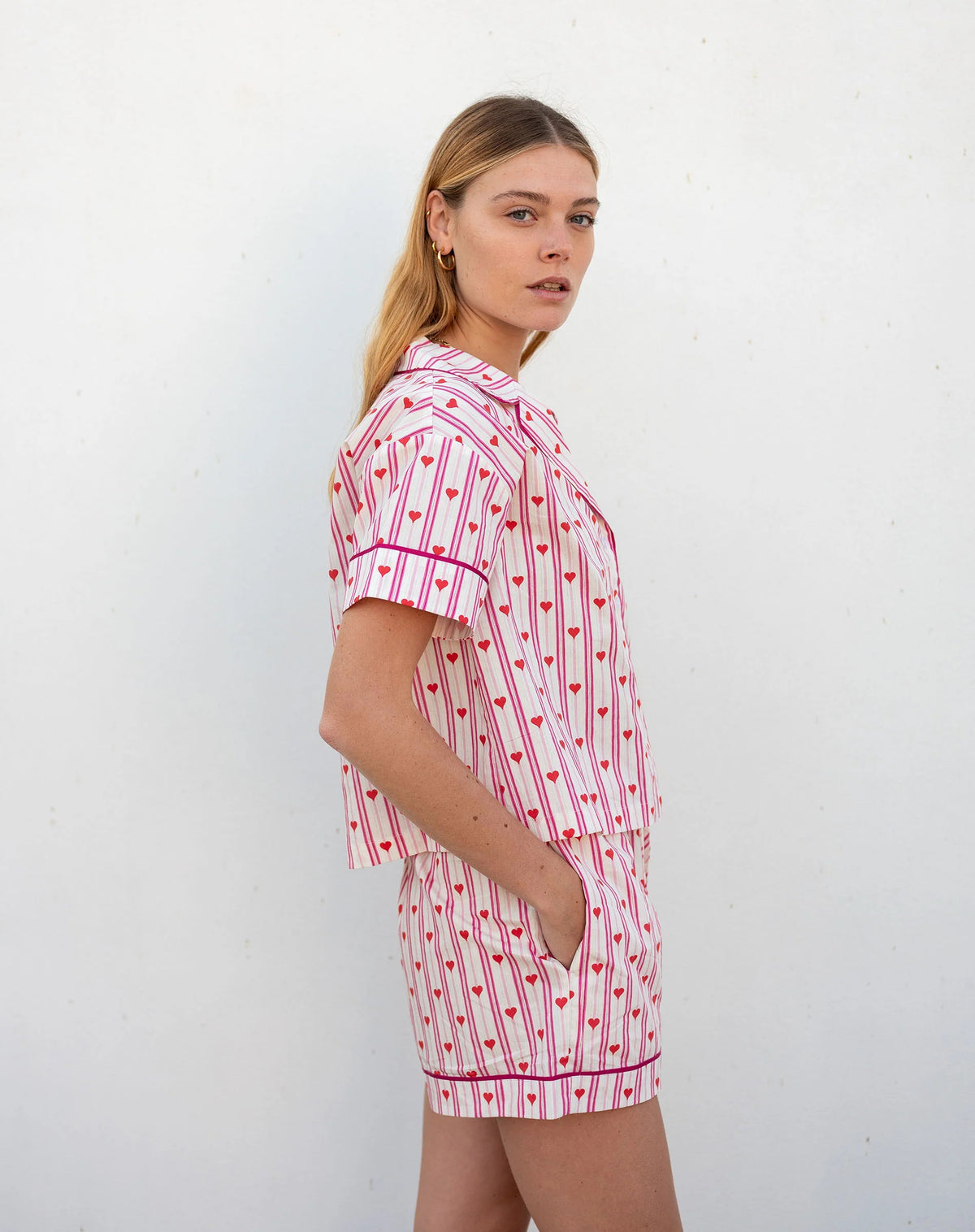 A woman with light brown hair smiles in MERSEA&#39;s OVER THE MOON PAJAMA SHORT SET LOVE NOTES— a heart print cotton short-sleeve button-up top and shorts—standing against a white background.