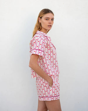 A woman with light brown hair smiles in MERSEA's OVER THE MOON PAJAMA SHORT SET LOVE NOTES— a heart print cotton short-sleeve button-up top and shorts—standing against a white background.
