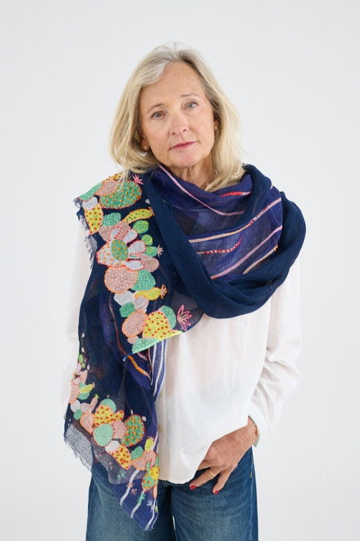 A blonde woman in a white blouse and blue jeans stands against a plain background, wearing the STORIATIPIC AZUR SCARF IN MARINE—100% linen with colorful handmade cactus embroidery.