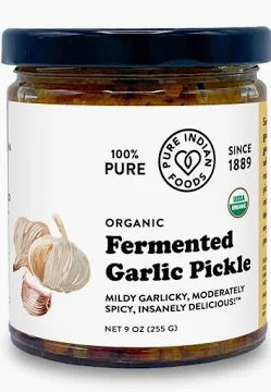 An open jar of PURE INDIAN FOODS-FERMENTED GARLIC PICKLE with a spoonful of chunky, reddish-orange fermented garlic held above it. The label notes it&#39;s mildly garlicky and moderately spicy. Brand: PURE INDIAN FOODS.