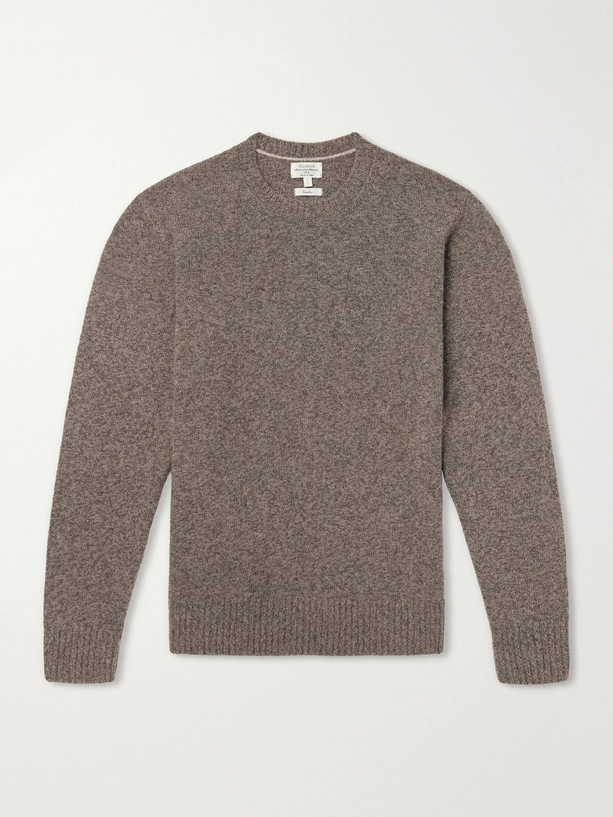 A person wearing the HARTFORD Seamless Wool Crew Pullover in Mushroom, an Italian-made sweater, paired with dark pants, is seated with one arm resting on their knee against a plain light background.