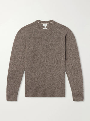A person wearing the HARTFORD Seamless Wool Crew Pullover in Mushroom, an Italian-made sweater, paired with dark pants, is seated with one arm resting on their knee against a plain light background.