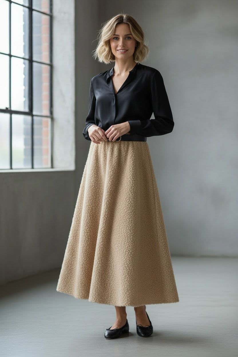 A woman stands indoors by large windows, smiling softly at the camera while wearing a black blouse, black flats, and the MIRTO 1956 TUILE CIRCLE SKIRT IN BEIGE by MIRTO 1956.