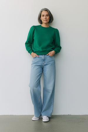 A woman with short gray hair stands against a plain wall, wearing the KMJ - COOPER SWEATSHIRT IN KELLY GREEN by KMJ, paired with light blue wide-leg jeans and white sneakers. She keeps her hands in her pockets and maintains a neutral expression.