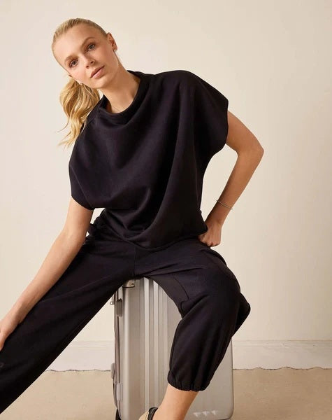 A woman with blonde hair in a ponytail wears the MERSEA Frenchie Dolman Top in Black and black joggers, sitting casually on a silver suitcase against a plain beige wall, effortlessly showcasing the relaxed silhouette of the MERSEA brand.