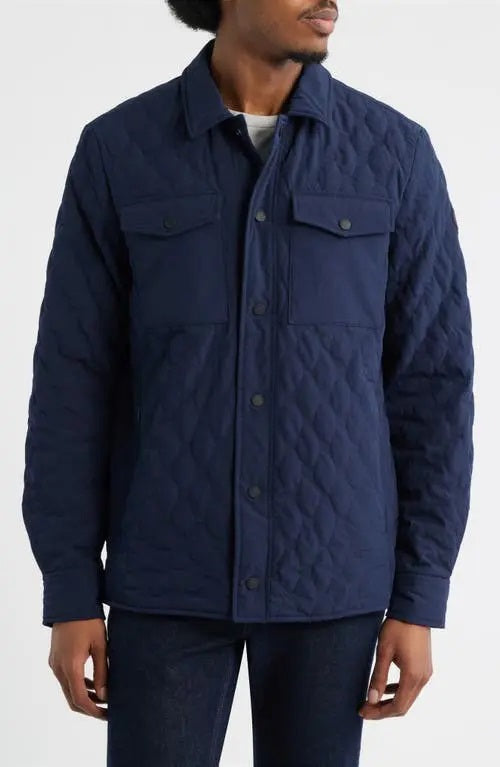 A man wears the SAVE THE DUCK Anton Jacket in navy—a dark blue, water-resistant jacket with buttoned chest pockets and a collar—paired with dark pants, standing against a plain white background.