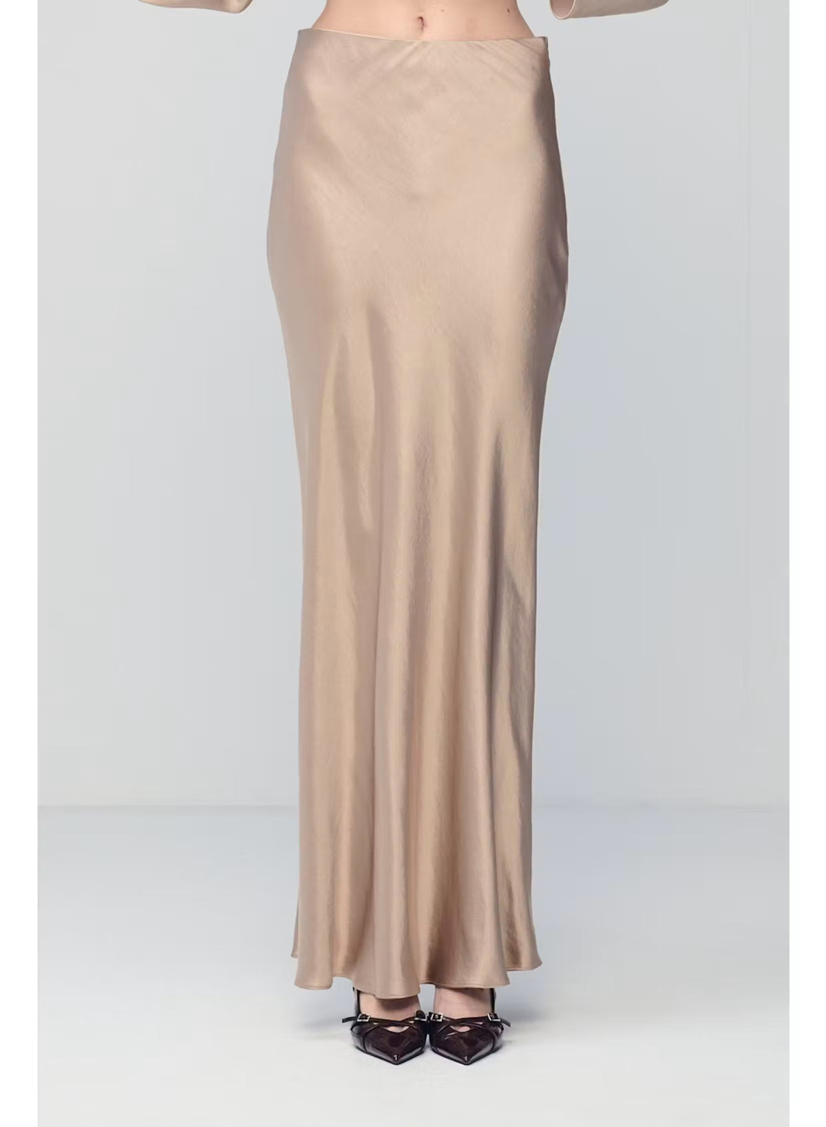 A person wearing the MILVA MI - LONG SATIN SKIRT IN SAND by MILVA, paired with pointed black shoes and standing against a plain, light background.