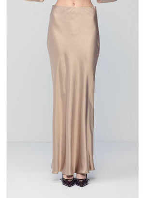 A person wearing the MILVA MI - LONG SATIN SKIRT IN SAND by MILVA, paired with pointed black shoes and standing against a plain, light background.