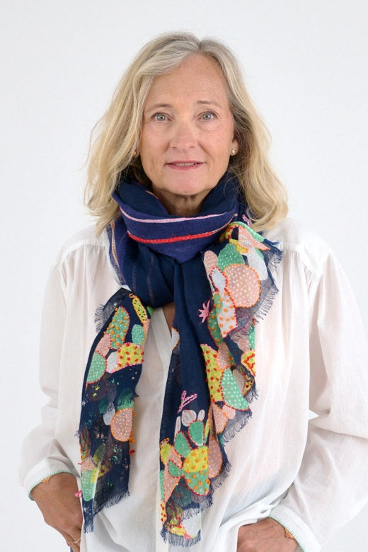 A blonde woman in a white blouse and blue jeans stands against a plain background, wearing the STORIATIPIC AZUR SCARF IN MARINE—100% linen with colorful handmade cactus embroidery.