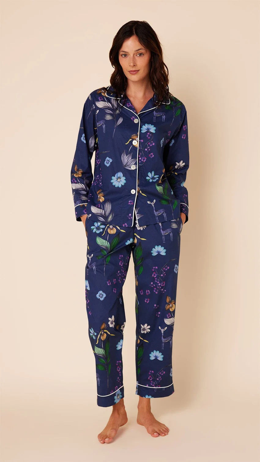A woman stands barefoot against a plain background, wearing the CAT&#39;S PAJAMAS - Deerly Luxe Pima Cotton Pajamas by CAT&#39;S PAJAMAS. The navy blue pajama set features a colorful floral pattern and is crafted from ultra-soft pima cotton. It includes a long-sleeved button-up shirt and matching pants, both adorned with white piping along the edges.