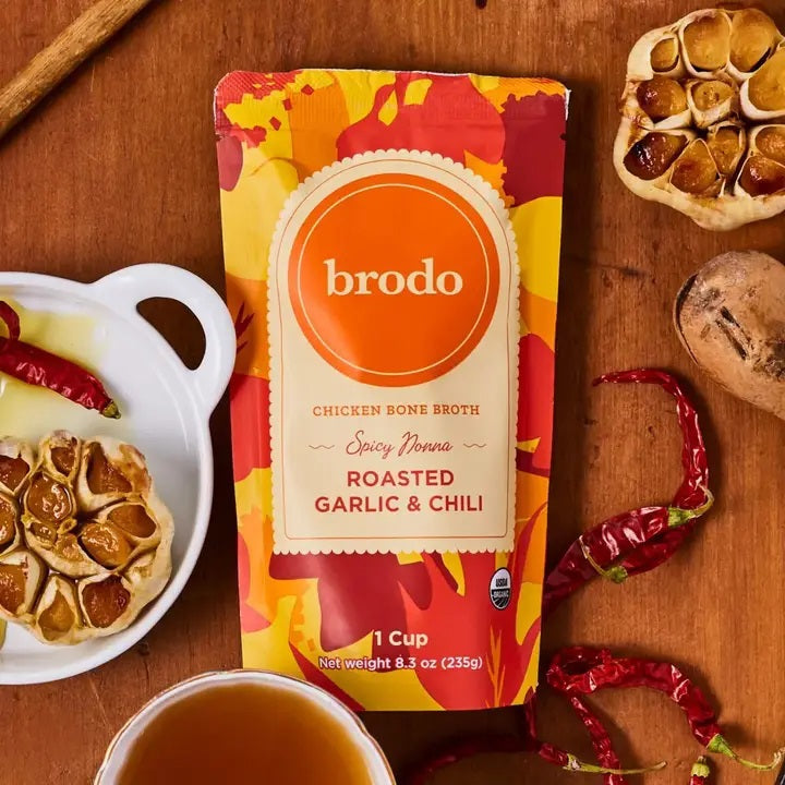 A vibrant package of BRODO BROTH COMPANY’s BRODO BROTH- SPICY NOONA ROASTED GARLIC & CHILI BONE BROTH ORGANIC rests on a wooden surface, surrounded by dried chili peppers, roasted garlic, and a steaming cup of flavorful chicken bone broth.