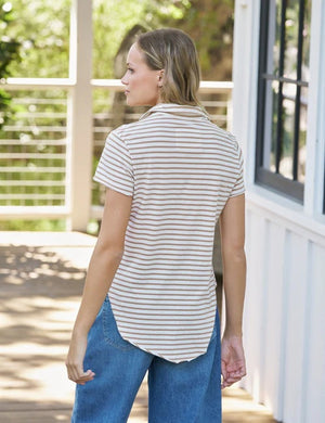 A woman with blonde hair stands outdoors, looking at the camera, wearing the FRANK & EILEEN Charlotte Perfect Polo in Heritage Jersey Wintage White and Camel Stripe. Greenery and white flowers are softly blurred in the background.