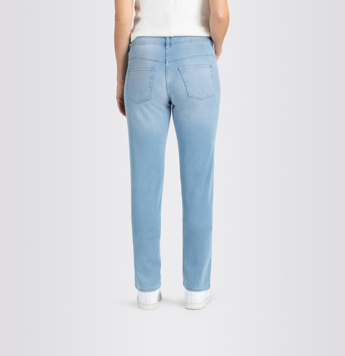 A person is standing against a plain background wearing MAC&#39;s DREAM STRAIGHT LEG DENIM jeans in light blue, paired with a white shirt and white sneakers. Their right hand is in their pocket while their left hand hangs down by their side. The slim fit of the MAC jeans provides a subtle shaping effect for a tailored look.