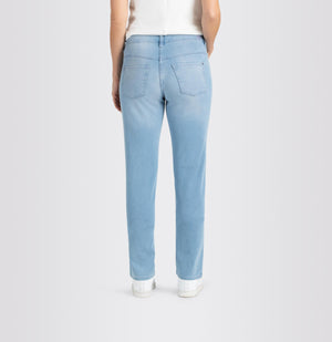 A person is standing against a plain background wearing MAC's DREAM STRAIGHT LEG DENIM jeans in light blue, paired with a white shirt and white sneakers. Their right hand is in their pocket while their left hand hangs down by their side. The slim fit of the MAC jeans provides a subtle shaping effect for a tailored look.