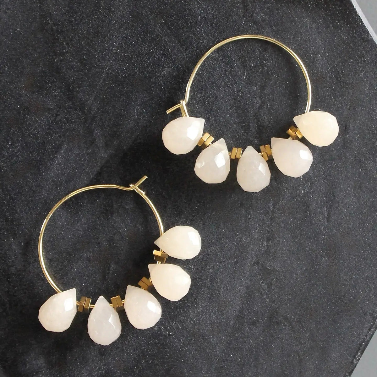 The DAVID AUBREY - DYED JADE MINI HOOP EARRINGS feature gold-plated hoops embellished with five teardrop-shaped, opaque white stones on each side, gracefully suspended from the hoops and displayed on a dark textured surface. Delicate gray jade beads from DAVID AUBREY accentuate the design, infusing these statement earrings with an elegant touch.