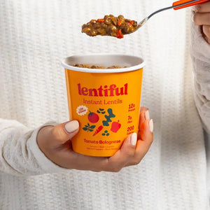 A container of LENTIFUL - LOW SODIUM TOMATO BOLOGNESE INSTANT LENTILS by LENTIFUL is surrounded by fresh carrots, spinach, tomatoes, bell pepper, beans, peas, herbs, and red pepper flakes for a heart-healthy meal.