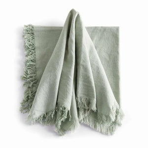 A stack of NAPA HOME AND GARDEN - AURA DINNER NAPKIN IN LIGHT GREEN, featuring fringed edges, is folded and placed on a white surface.