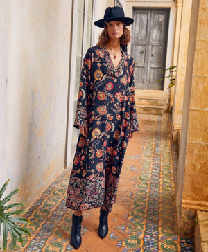 The MOMONI - FELICIEN DRESS by MOMONI is a long-sleeved, ankle-length black crepe de chine dress with a vibrant floral and paisley print in red, orange, and green, finished with patterned borders at the hem and cuffs.