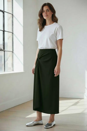 A woman stands indoors in natural light, wearing a white T-shirt paired with the MEIMEIJ Midi Skirt with Side Gather in dark green from MEIMEIJ, styled with silver flats. She has wavy brown hair and a neutral expression.