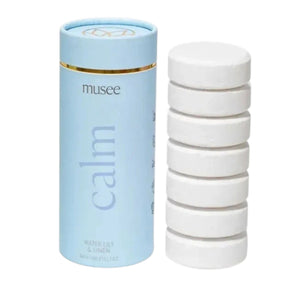 A person in striped pajamas holds three pastel-colored containers labeled “MUSEE,” including one for the MUSEE - CALM BATH TABLETS. These natural bath products by MUSEE, designed to promote a soothing soak, come in yellow-green, blue, and lavender hues.