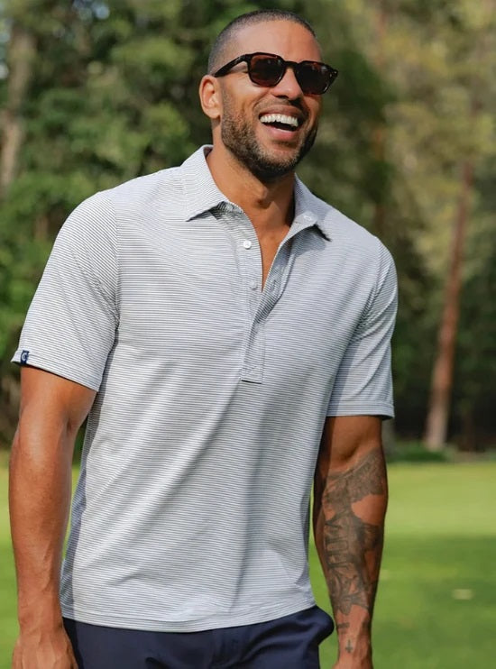 A smiling man with short hair and a beard wears the CRIQUET - Performance Sport Range Polo in Hobby Stripe - Gray as he holds a canned drink in a green koozie, facing slightly to the left against a plain white background.