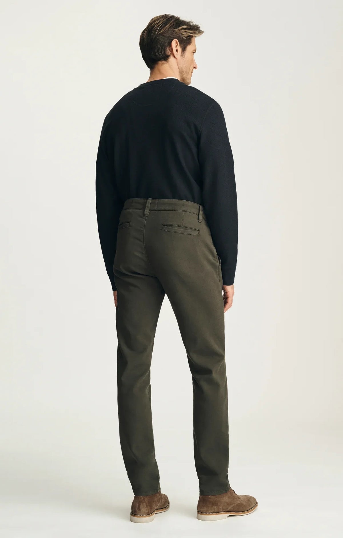 A person stands with their back to the camera against a neutral background, wearing the 34 HERITAGE - VERONA Straight Leg Chino Pant in Jasper High Flyer paired with a black long-sleeve shirt and brown suede shoes.