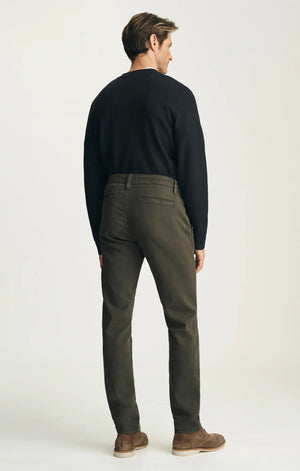 A person stands with their back to the camera against a neutral background, wearing the 34 HERITAGE - VERONA Straight Leg Chino Pant in Jasper High Flyer paired with a black long-sleeve shirt and brown suede shoes.