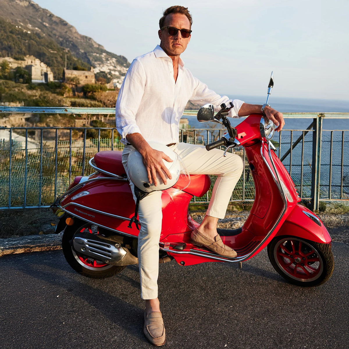 Wearing a COLLARS AND CO - TOSCANO LINEN SHIRT IN WHITE, a man with sunglasses sits on a red scooter by a scenic coastal road, holding a white helmet, with hills, buildings, and the sea in the background.