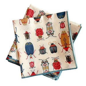 The BEETLE COCKTAIL NAPKIN by DOT AND ARMY features a playful red, blue, and yellow beetle and bug pattern on soft white cotton. Perfect for adding a fun touch to your table setting. Two napkins shown on a light wooden surface.