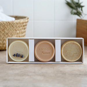 A hand holds a L'AUGUSTE PROVENCE handcrafted French soap bar and rubs it on a person's leg, creating a creamy lather that suggests an exfoliating skincare routine using artisanal soaps from L'AUGUSTE PROVENCE.