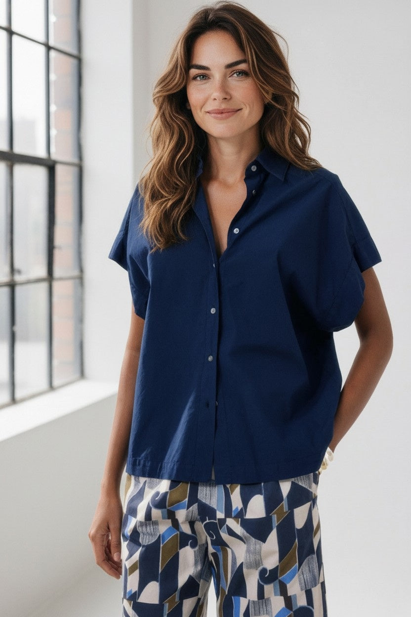 A woman with long brown hair, wearing the HARTFORD - THE CARLOS SHIRT IN NIGHT by HARTFORD and patterned pants, stands indoors by a large window, smiling softly.