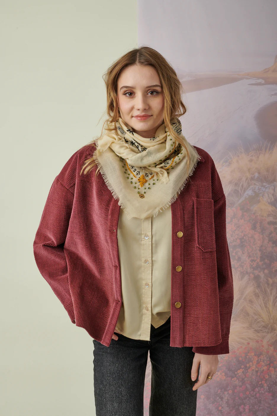 A young woman with light brown hair wears the STORIATIPIC Nelly Square Cashmere and Wool Embroidered Bandana Scarf, paired with a light gray top, standing before a softly colored nature-themed backdrop.