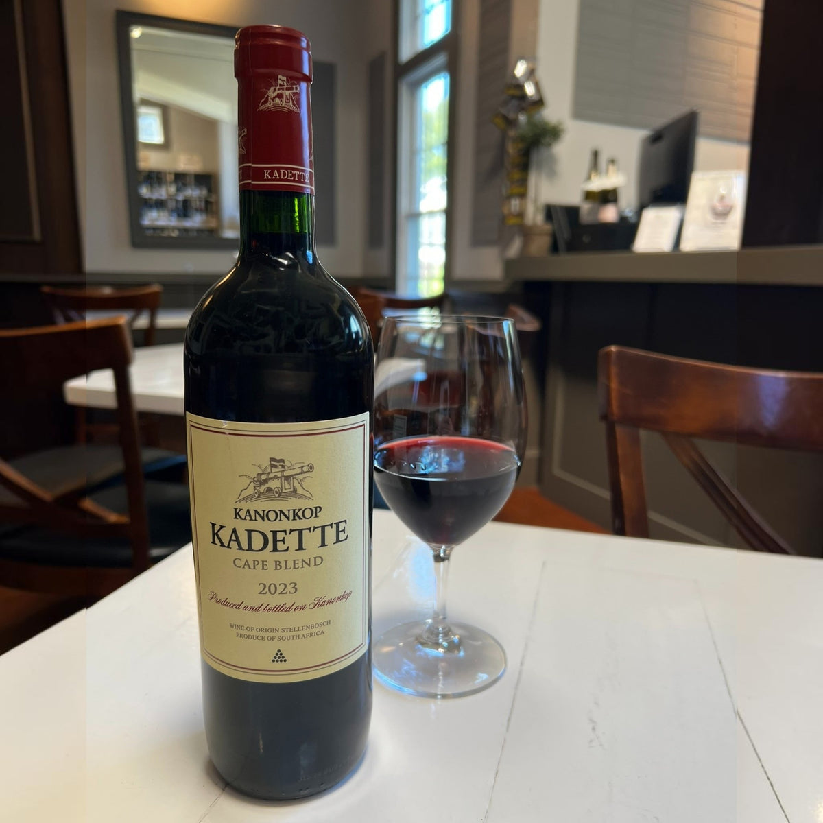 A bottle of KANONKOP ‘ KADETTE CAPE BLEND’ 2023 by WINEBOW, featuring a cream label and red cap, stands upright against a white background.