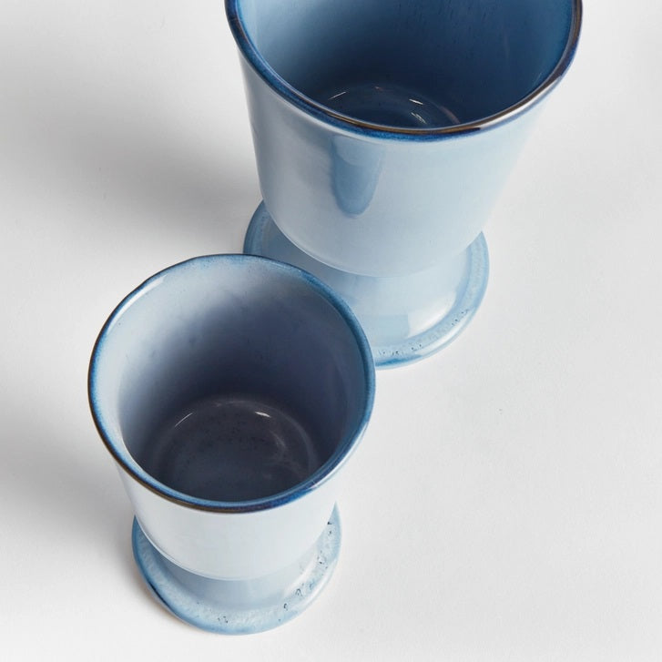 Two NAPA HOME AND GARDEN Blue Kelcy Footed Cachepot planters sit on a wooden table with a blue and white striped placemat; one holds a small leafy green plant, the other is empty. A white textured wall is in the background.
