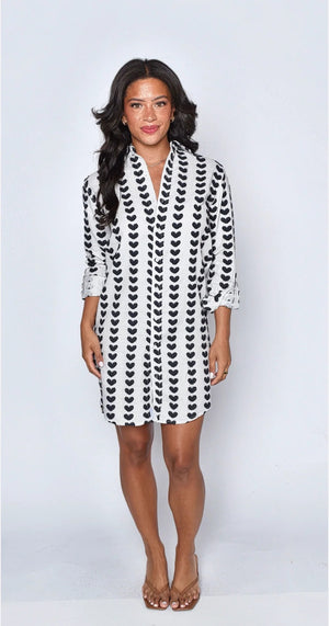 A woman stands indoors near a large window, smiling as she wears the EMILY PHILLIPS - THE BLACK HEARTS MINI SHIRTDRESS—a versatile white long-sleeve piece featuring black heart patterns—paired with blue jeans and black ankle boots.