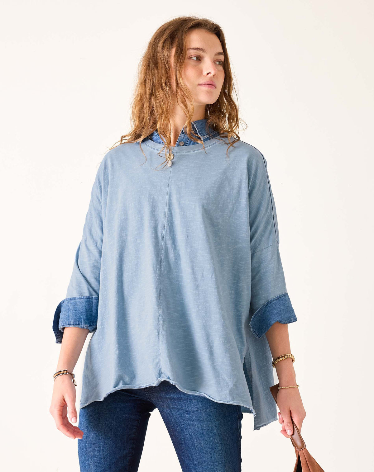 A woman with wavy light brown hair wears the MERSEA - Catalina Slub Tee in Shore Blue Stitches with rolled-up sleeves over denim jeans, looking to her left against a plain, light background.