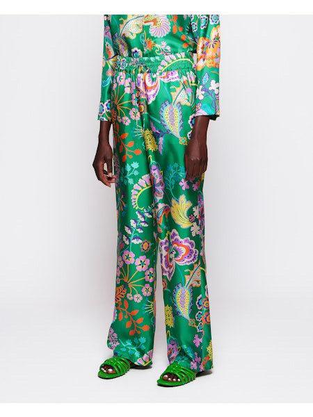 A person in MIRTO 1956&#39;s silk floral pant, featuring an elastic waistband, holds a large striped tote bag. Complementing the outfit are matching green sandals, all set against a plain white background.