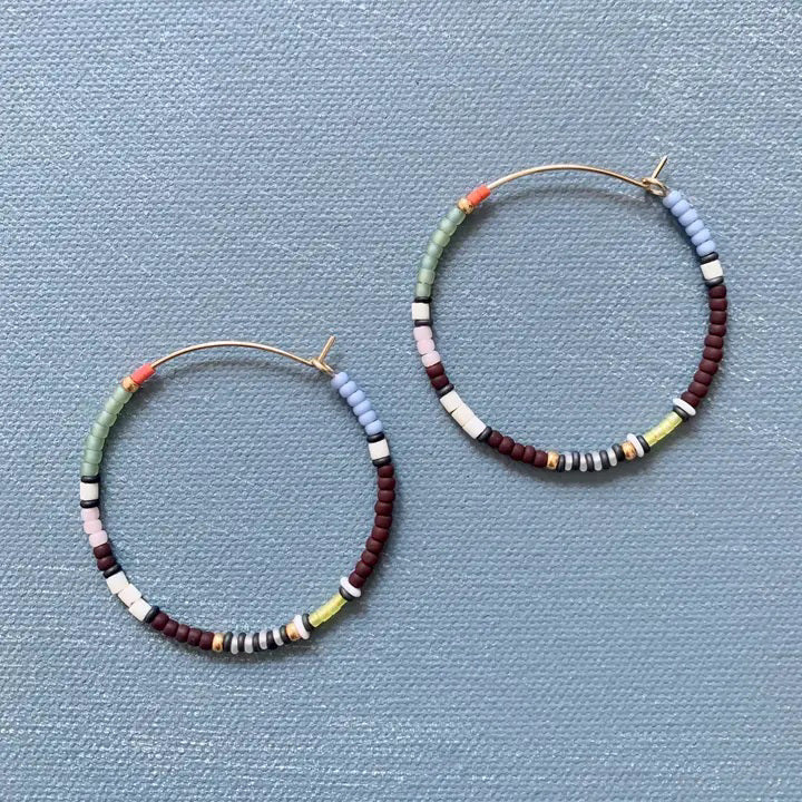 The ALICE RISE - BIG COLORLOOP EARRINGS IN LICORICE by ALICE RISE, handmade in North Carolina and featuring vibrant multicolor beads, are displayed on a textured light blue background.