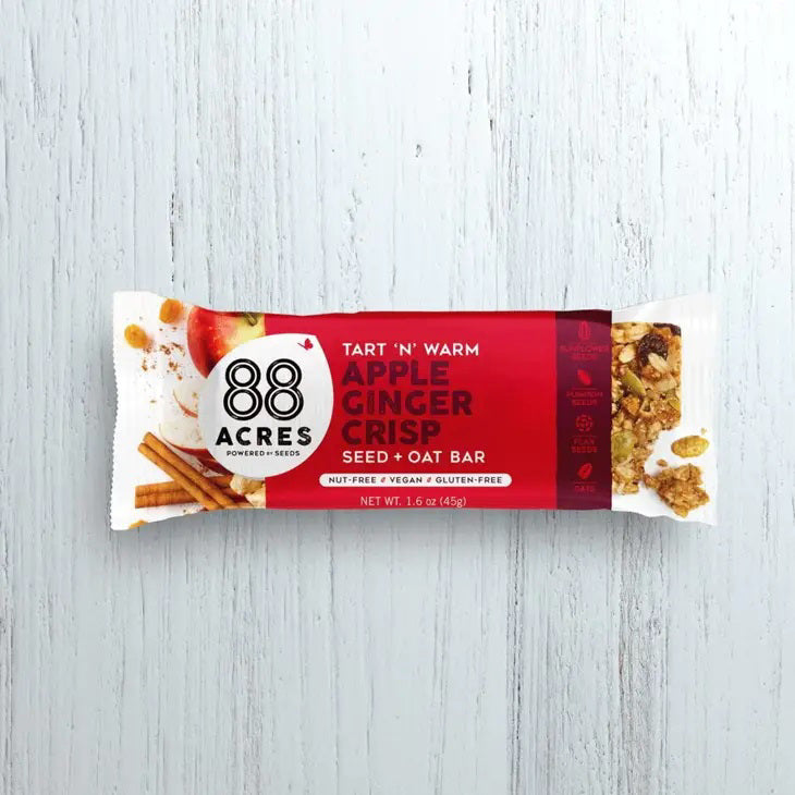 A package of 88 ACRES - APPLE GINGER CRISP SEED + OAT BAR rests on a light wooden surface. The primarily white and red wrapper emphasizes its nut-free, vegan, and gluten-free benefits. The bar is partially visible on the right side.