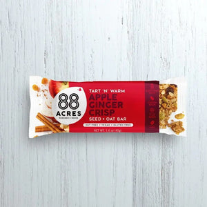 A package of 88 ACRES - APPLE GINGER CRISP SEED + OAT BAR rests on a light wooden surface. The primarily white and red wrapper emphasizes its nut-free, vegan, and gluten-free benefits. The bar is partially visible on the right side.